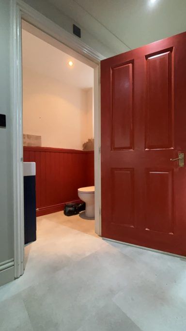 Interior view of a bathroom with a red door and light-coloured walls, featuring a toilet and sink.