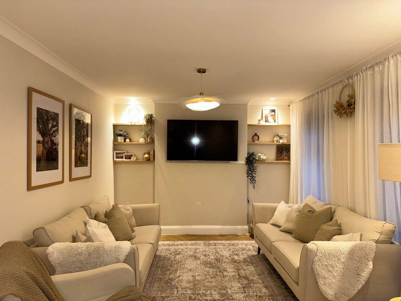 A cosy living room with a TV, two sofas, shelves, and soft lighting.