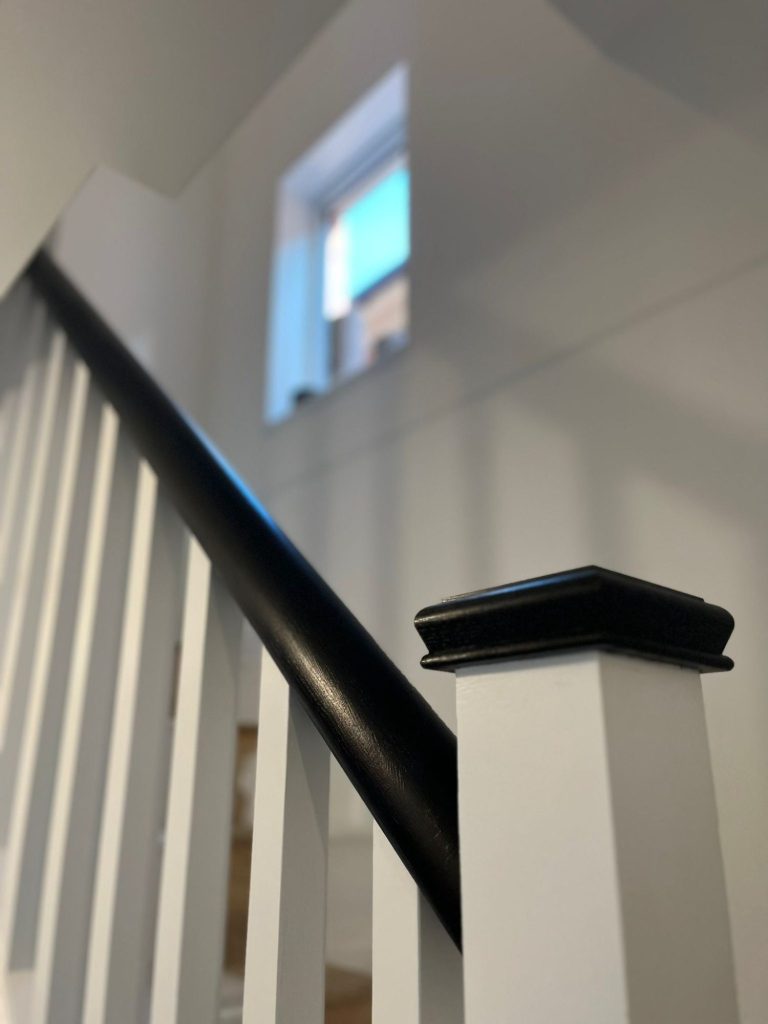 Black handrail and white wooden staircase leading to a window with a glimpse of blue sky.