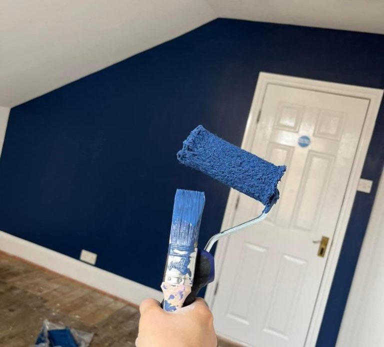 Hand holding paint rollers in a room with a recently painted dark blue wall.