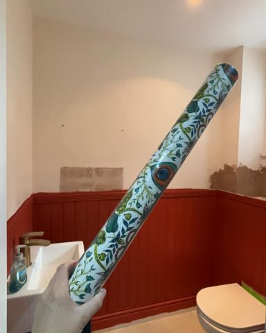 A hand holding a roll of patterned wallpaper in a bathroom setting.